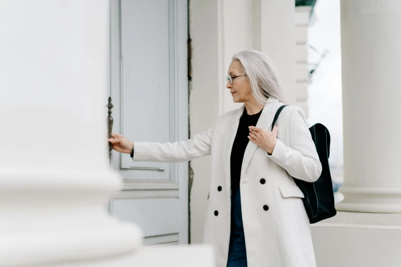 a woman in a white coat standing in front of a building, pexels contest winner, opening door, silver haired, background image, people looking at a house