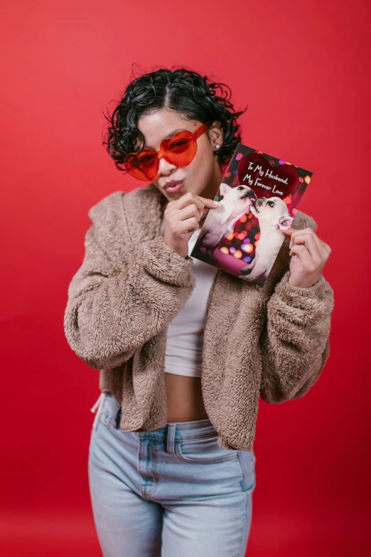 a woman holding a magazine in front of her face, an album cover, by Julia Pishtar, pop art, red sunglasses, furr covering her chest, with a cup of hot chocolate, official store photo