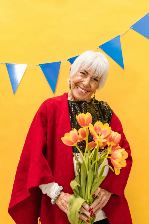 an older woman holding a bouquet of tulips, an album cover, yellow robes, bright white hair, happy and spirited expression, portrait photo of a backdrop