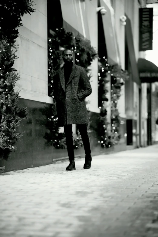 a black and white photo of a man standing on a sidewalk, inspired by Peter Lindbergh, harlem renaissance, wearing festive clothing, gif, ( ( wearing a long coat ) ), brown skinned