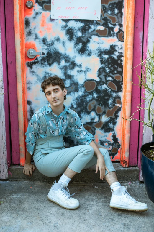 a man sitting in front of a door next to a potted plant, an album cover, by Jessie Alexandra Dick, trending on pexels, graffiti, attractive androgynous humanoid, wearing a light blue shirt, natalia dyer, patterned clothing