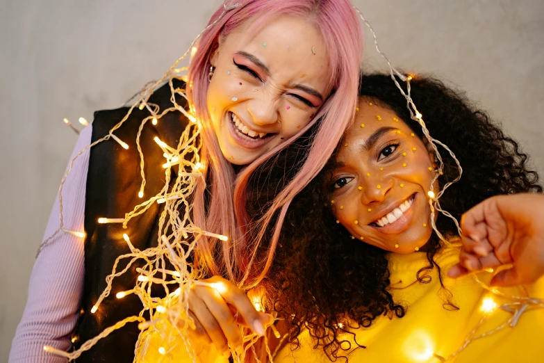 a couple of women standing next to each other, trending on pexels, happening, fairy lights, pink and yellow, with gold teeth, diverse