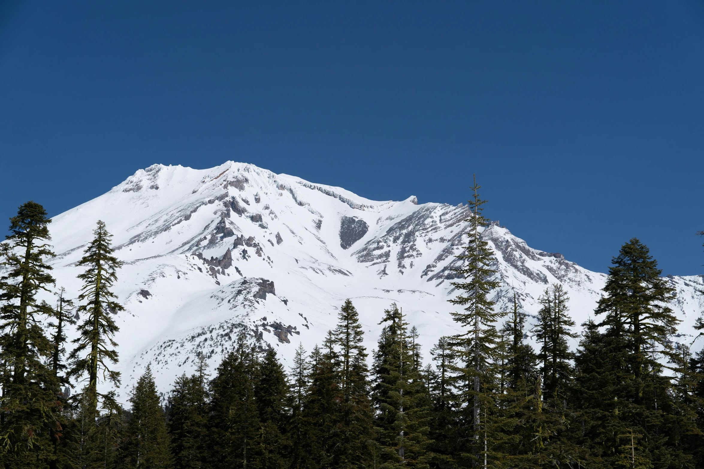 a man riding skis down a snow covered slope, a photo, giant crater in distance, clear blue skies, majestic forest grove, with snow on its peak