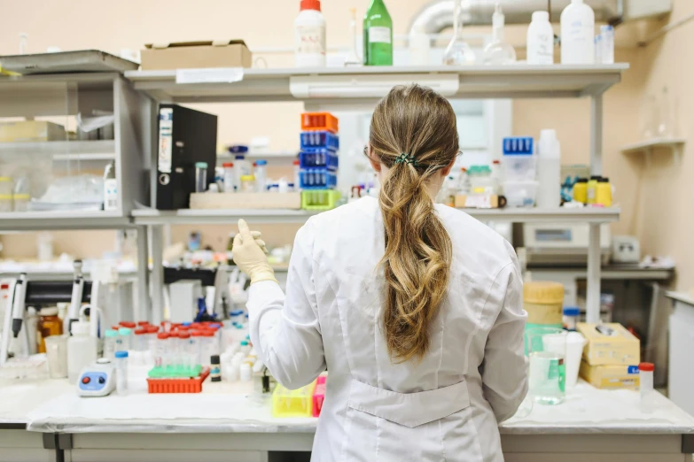 a woman in a lab coat standing in front of a counter, by Meredith Dillman, pexels, long shot from the back, sterile colours, college, engineering