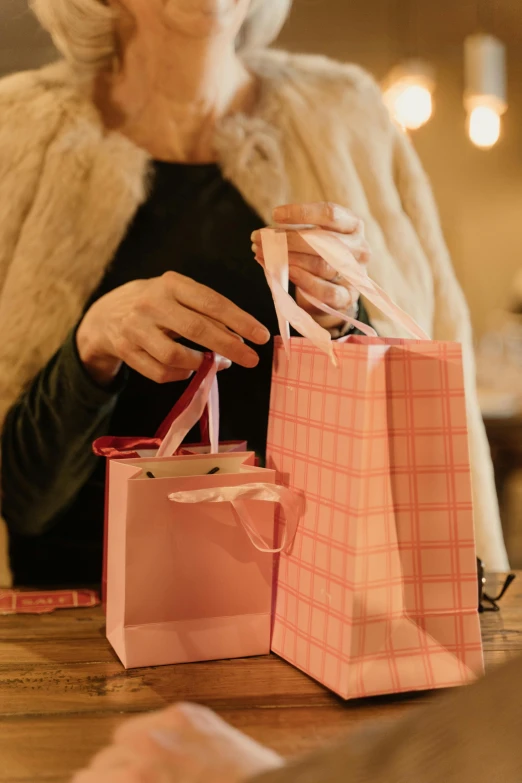 a woman sitting at a table with shopping bags, by Helen Stevenson, pexels, pink and pink details, giving gifts to people, softly lit, ribbon