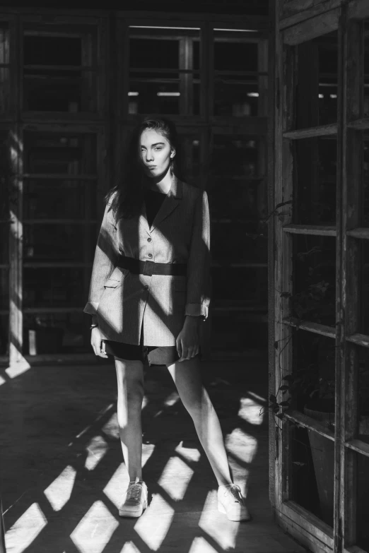 a black and white photo of a woman standing in a doorway, inspired by Peter Lindbergh, bauhaus, the girl and the sun, hybrid of gal gadot, short skirt and a long jacket, brutalist fashion show