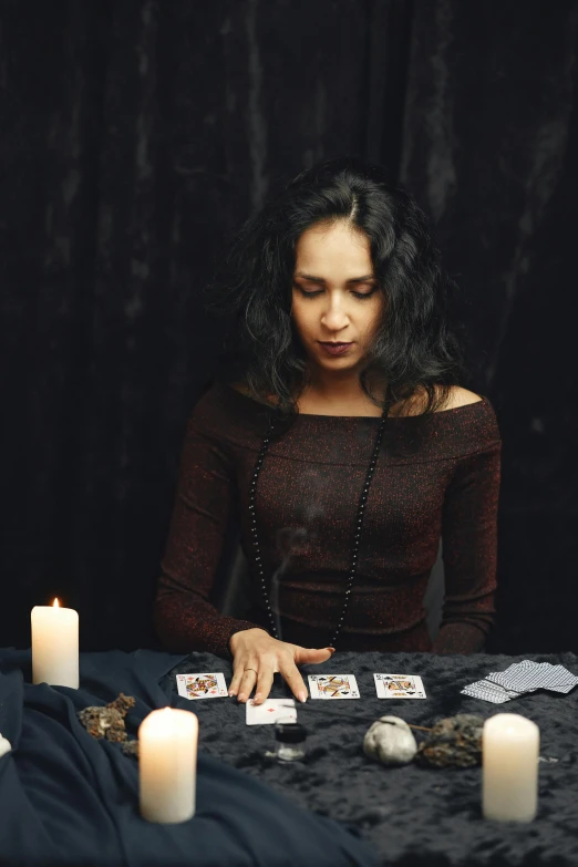 a woman sitting at a table with candles and playing cards, an album cover, trending on pexels, woman in black robes, black - haired mage, 2 0 0 0's photo, spell casting