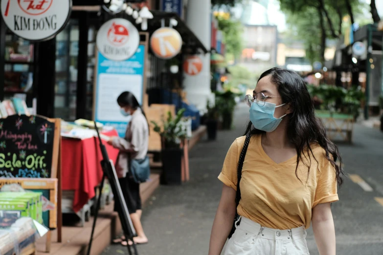 a woman wearing a face mask walking down a street, pexels, happening, south east asian with round face, avatar image, standing in a restaurant, student