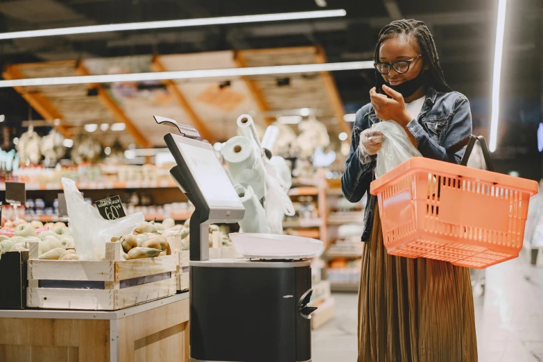 a woman standing in a grocery store looking at her cell phone, pexels contest winner, dark-skinned, 🦩🪐🐞👩🏻🦳, cash register, getting groceries