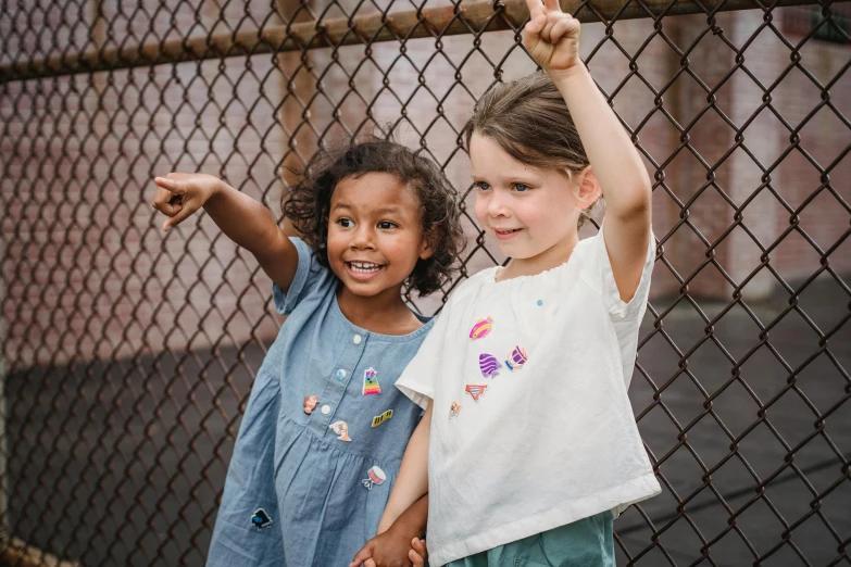 two little girls standing next to each other near a fence, a portrait, by Nick Fudge, pexels contest winner, embroidered shirt, playful and cheerful, varying ethnicities, stick poke