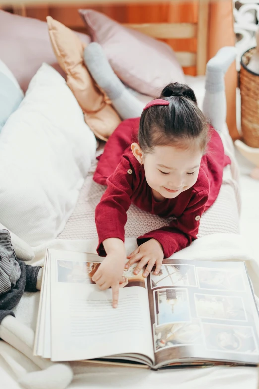 a little girl laying on a bed reading a book, a picture, inspired by Li Di, pexels contest winner, crimson themed, old photobook, inspect in inventory image, young asian girl