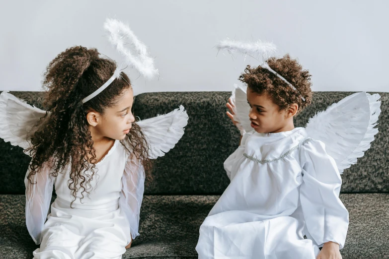 two little girls dressed as angels sitting on a couch, by Marie Angel, pexels contest winner, varying ethnicities, feather hair ornaments, biblically acurate angel, thumbnail