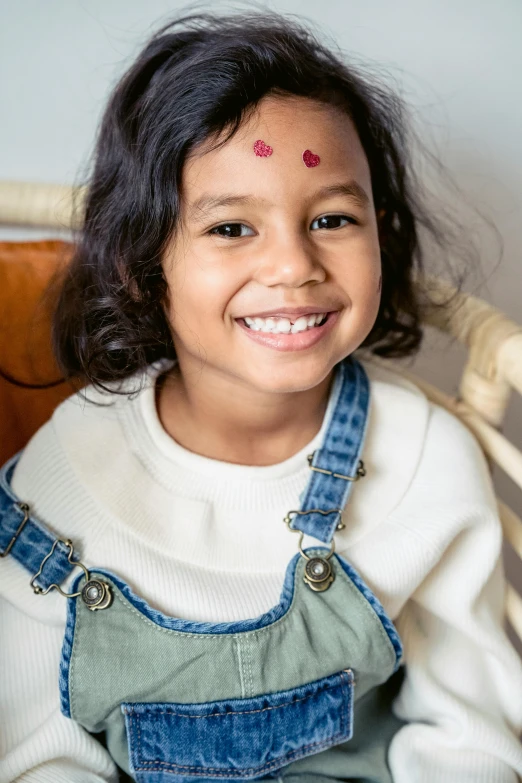 a little girl sitting on top of a wooden chair, trending on pexels, jeweled ornament over forehead, subject is smiling, wearing a patch over one eye, south east asian with round face