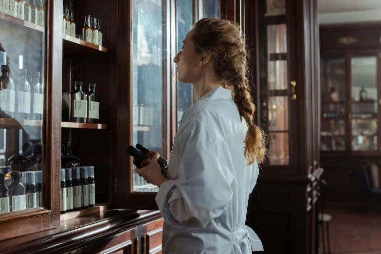 a woman standing in front of a cabinet holding a cell phone, by Emma Andijewska, pexels contest winner, apothecary, standing in a cantina, profile image, wearing a labcoat