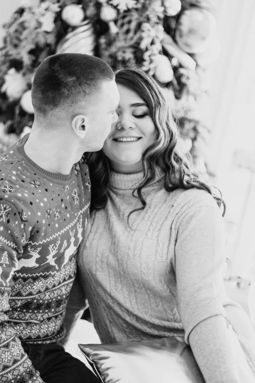 a man and woman sitting in front of a christmas tree, a black and white photo, by Julia Pishtar, kissing smile, wearing a sweater, andrey surnov, 🌸 🌼 💮