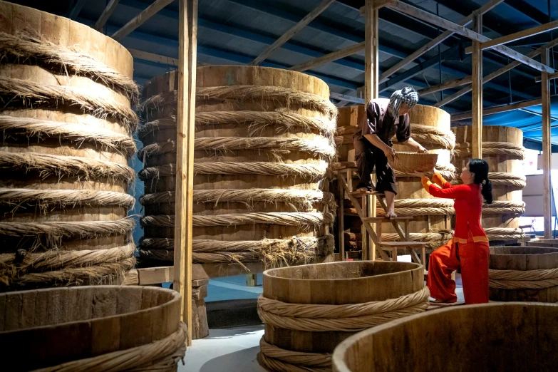 a woman standing next to a pile of wooden barrels, inspired by Fu Baoshi, process art, inside the building, rope bridges, avatar image, introduction factory photo