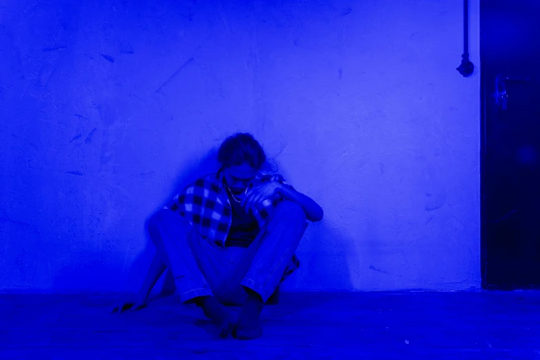 a person sitting on the ground in a dark room, blue backdrop, concerned, bright blue, ignant