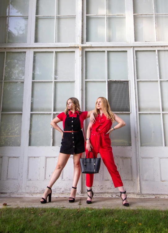 two women standing next to each other in front of a building, an album cover, trending on pexels, red jumpsuit, black shirt with red suspenders, 5 0 0 px models, in front of white back drop