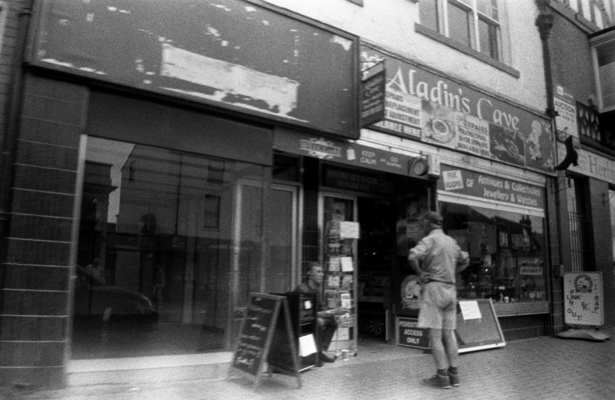 a black and white photo of a man standing in front of a store, a black and white photo, inspired by Bert Hardy, flickr, coventry city centre, disposable film, a busy arcade, magic shop