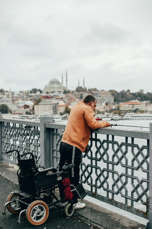 a man standing on top of a bridge next to a river, by Ismail Acar, trending on pexels, hurufiyya, sitting in a wheelchair, istanbul, gray men, 🚿🗝📝