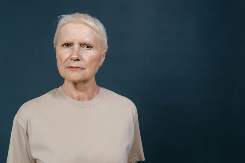 an older woman standing in front of a blue background, unsplash, hyperrealism, human staring blankly ahead, on a gray background, face and body clearly visible, looking serious