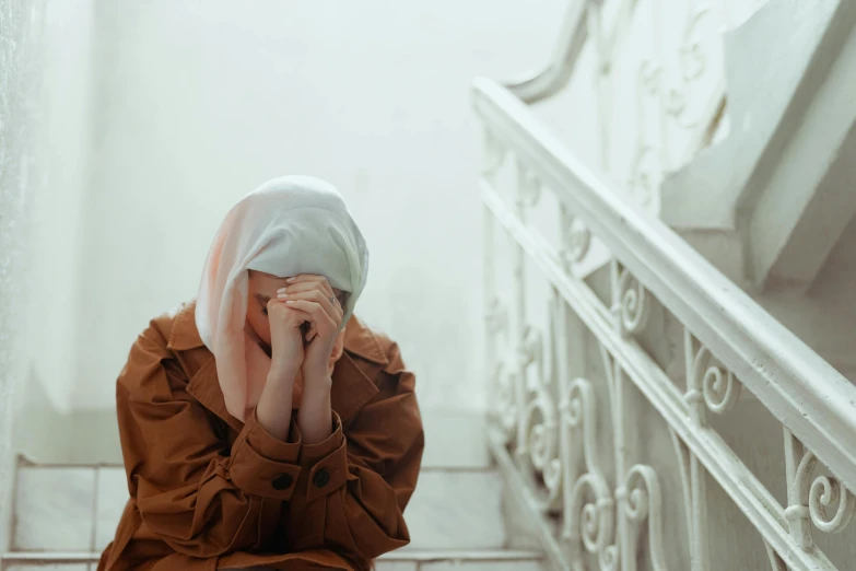 a woman sitting on the floor covering her face with a towel, trending on pexels, hurufiyya, white hijab, sit on a bench, distraught, brown