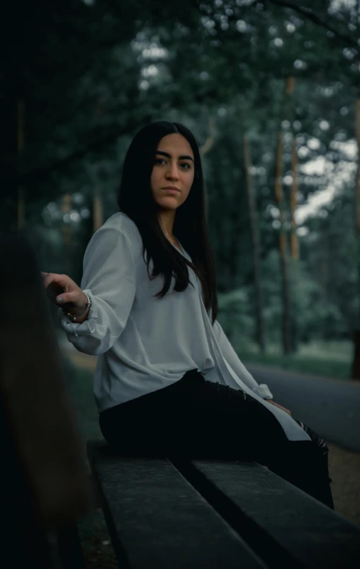 a woman sitting on a bench in a park, by Attila Meszlenyi, pexels contest winner, realism, wearing a white shirt, threatening pose, young middle eastern woman, standing in a dark forest