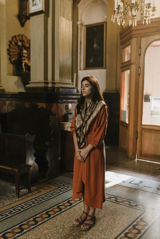 a woman standing in a room with a chandelier, inspired by Modest Urgell, pexels contest winner, qajar art, wearing brown robes, scene set in a church, in the style wes anderson, mia khalifa