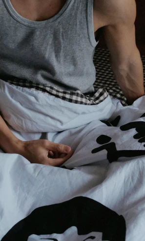 a man sitting on top of a bed covered in a blanket, a cartoon, inspired by Sesshū Tōyō, trending on pexels, closeup of arms, kanji markings, wearing a t-shirt, holding hand