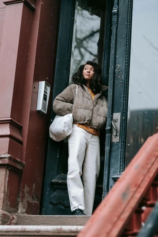 a woman standing on the steps of a building, by Nina Hamnett, trending on pexels, renaissance, model wears a puffer jacket, brown and white color scheme, brooklyn, exiting store