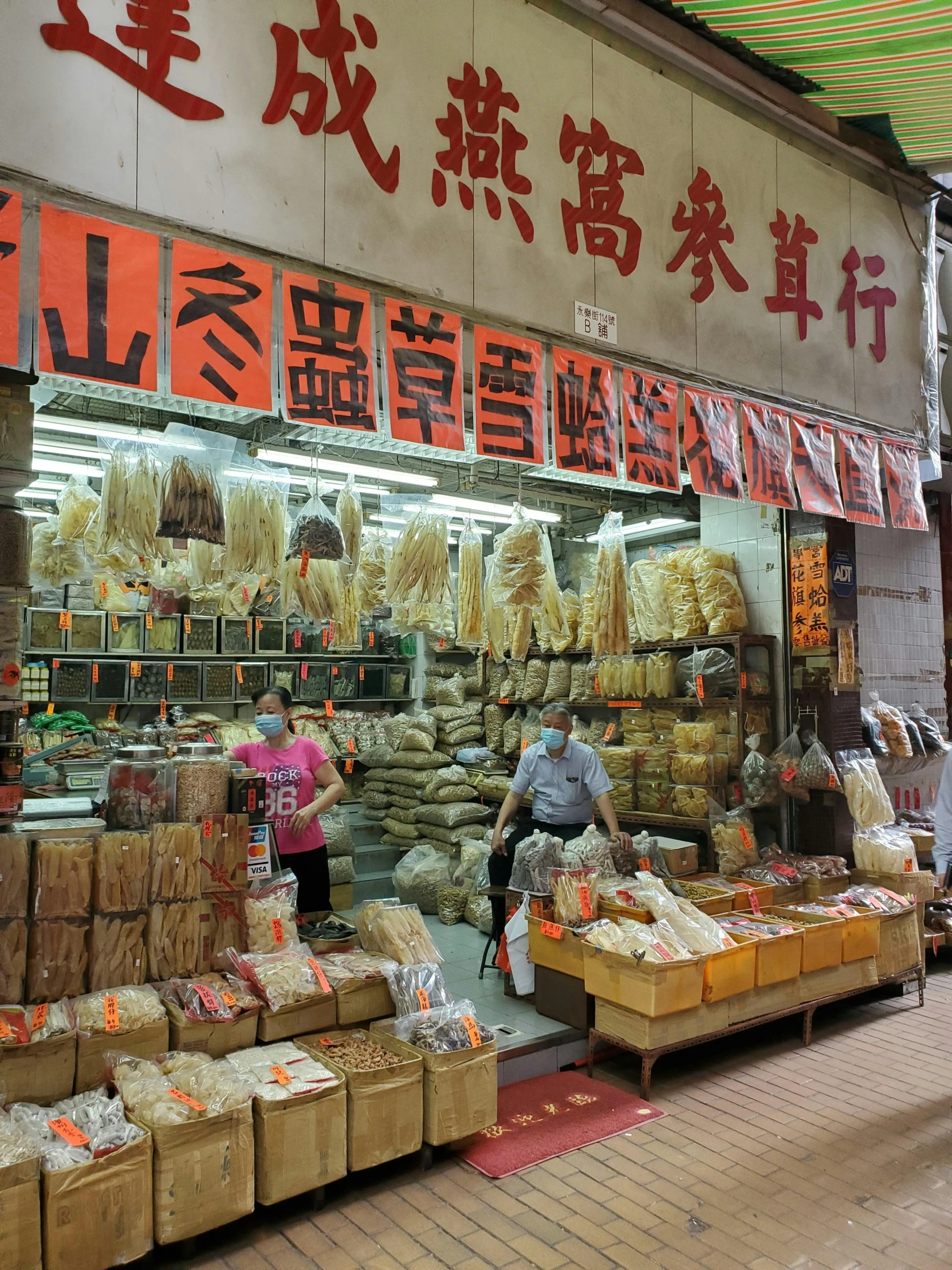 a group of people standing in front of a store, spices, hong kong buildings, dried plants, bags on ground