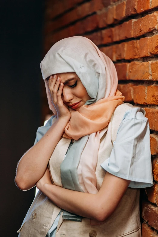 a woman in a hijab leaning against a brick wall, a picture, by Julia Pishtar, shutterstock, hurufiyya, stressed and burnt out, pastel', resting head on hands, wearing a scarf