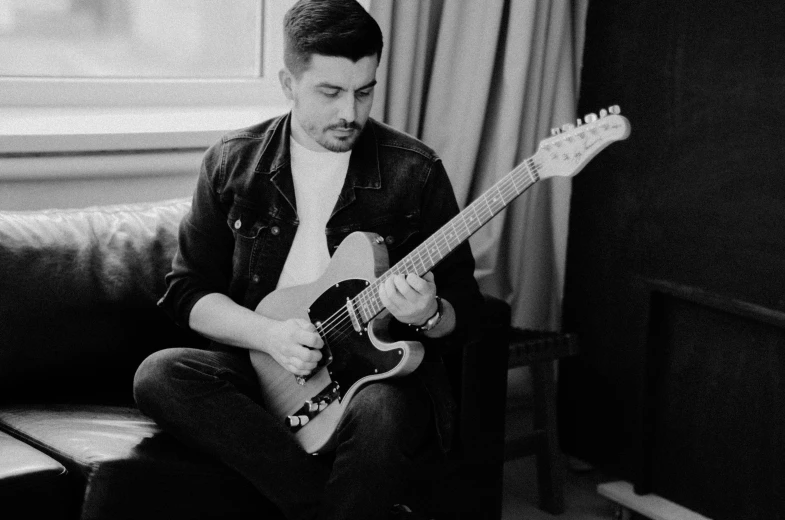 a man sitting on a couch playing a guitar, by Nick Fudge, tom burke, on a pale background, monochrome, lead guitar