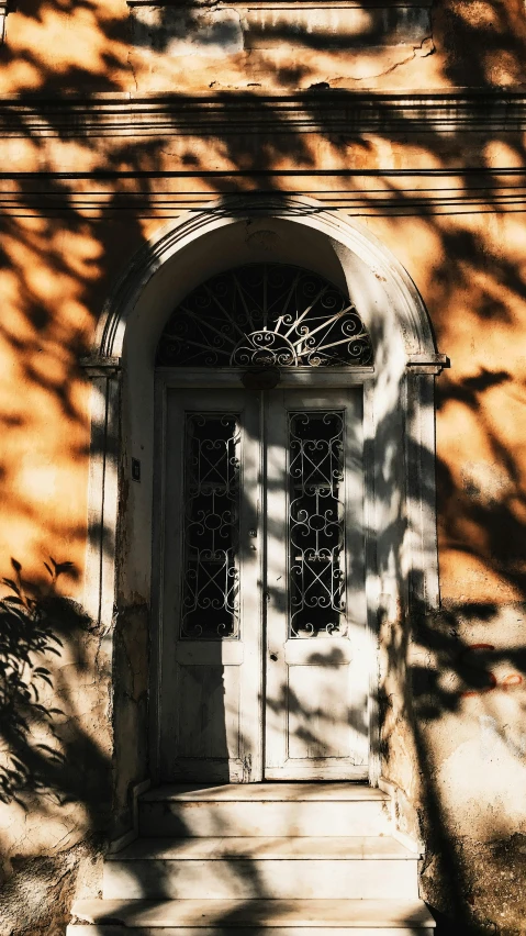 a white door sitting on the side of a building, by Constantine Andreou, pexels contest winner, neoclassicism, large tree casting shadow, warm glow, an overgrown, black and terracotta