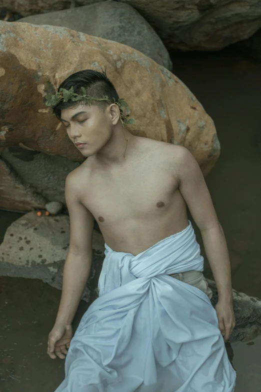 a man sitting on top of a rock next to a body of water, an album cover, inspired by Byron Galvez, sumatraism, wearing toga, nonbinary model, trending photo, dressed in laurel wreath