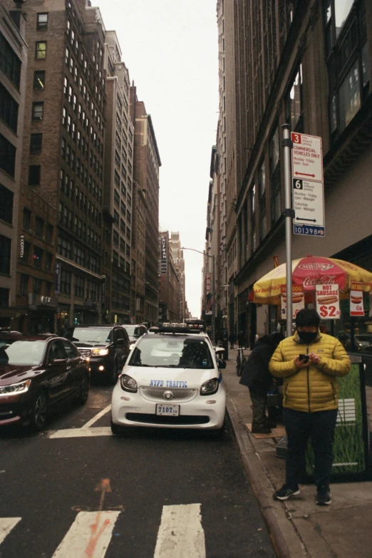 a white car driving down a street next to tall buildings, a picture, happening, humans of new york, smartphone photo, people, cars parked