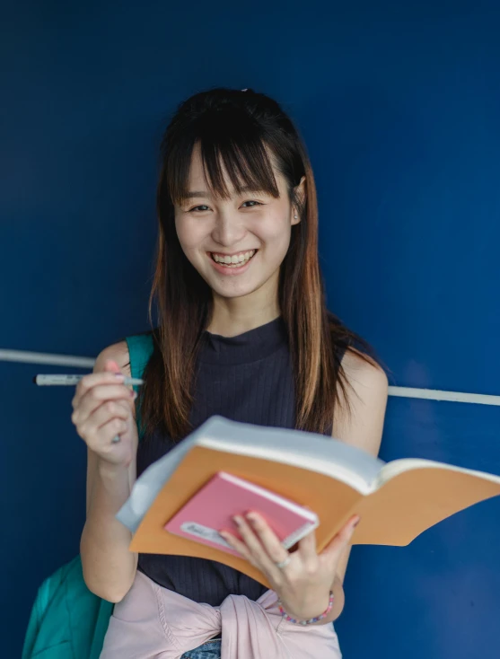 a woman standing in front of a blue wall holding a book, inspired by helen huang, pexels contest winner, academic art, smiling girl, ethnicity : japanese, center parted bangs, holding a stuff