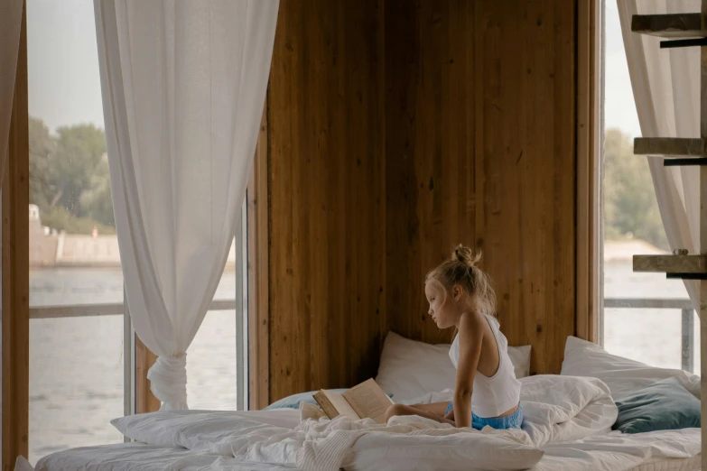 a little girl sitting on a bed reading a book, by Julia Pishtar, pexels contest winner, inside of a cabin, white sheets, in a beachfront environment, gif