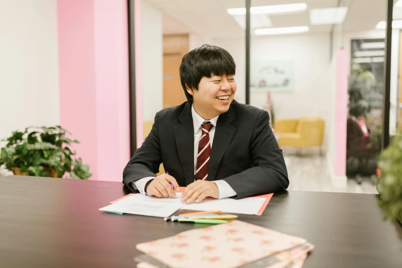 a man in a suit sitting at a table, by Jang Seung-eop, friendly smile, student, wearing a light - pink suit, for junior