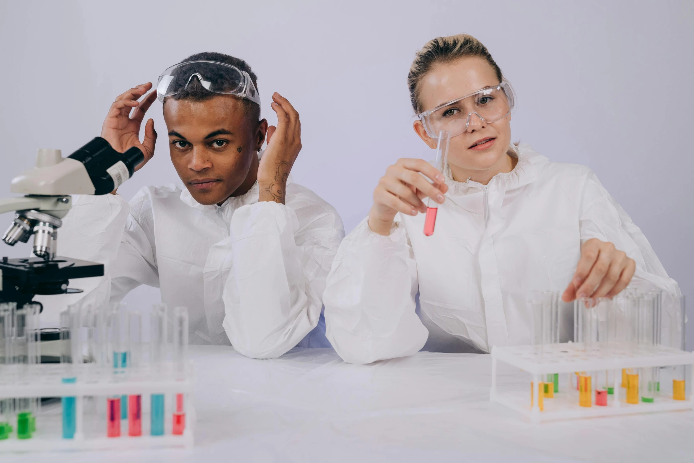 a couple of people sitting at a table in front of a microscope, trending on pexels, antipodeans, beakers of colored liquid, white robes, goggles on forehead, school class