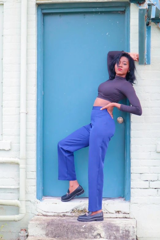 a woman standing in front of a blue door, an album cover, by Dulah Marie Evans, pexels contest winner, renaissance, blue pants, pose model, ( ( dark skin ) ), purple