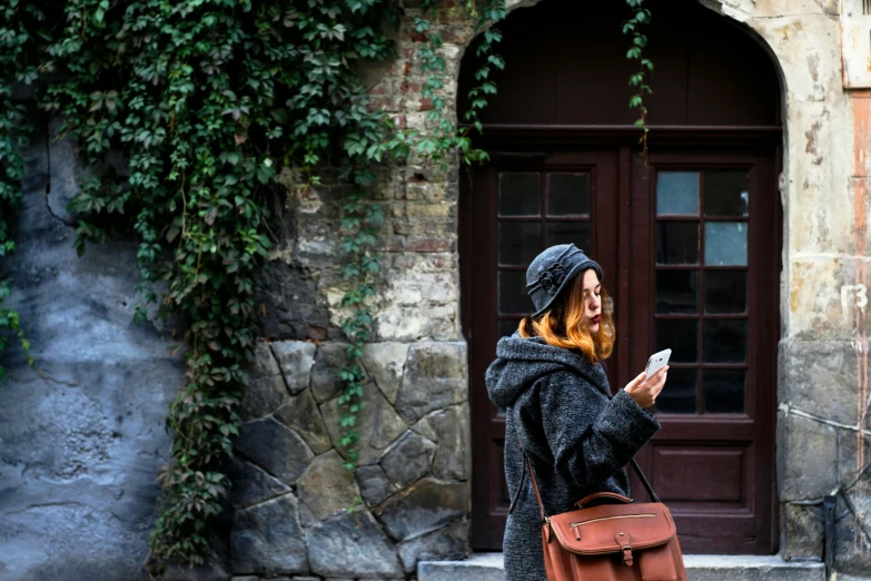 a woman standing in front of a building looking at her cell phone, trending on pexels, visual art, caracter with brown hat, outside the'school of magic ', ivy, a cozy