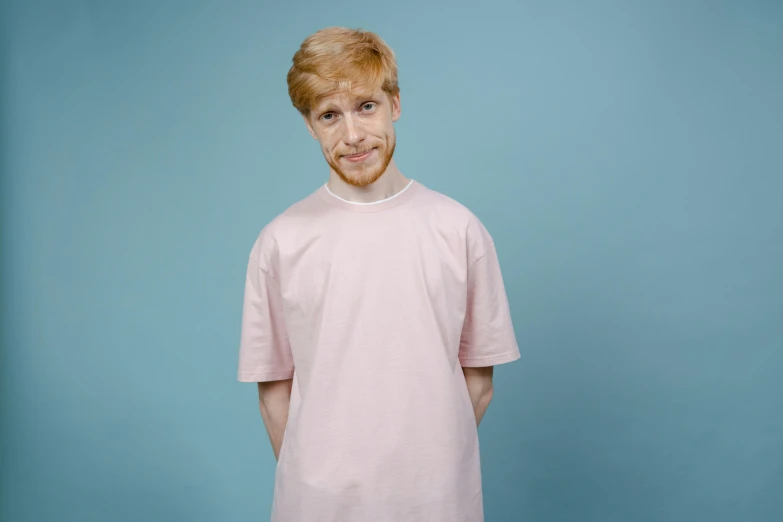 a young man standing in front of a blue background, an album cover, hyperrealism, ginger hair, pink shirt, wearing a hospital gown, on grey background