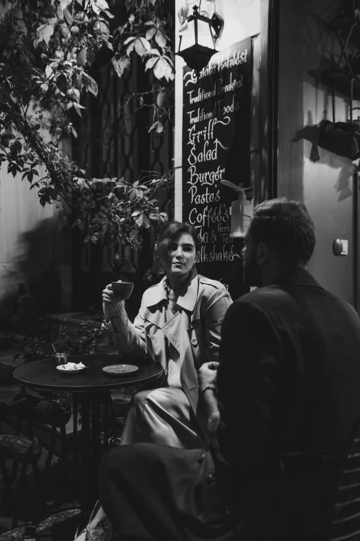 a black and white photo of two people sitting at a table, inspired by Brassaï, pexels contest winner, woman is in a trenchcoat, cozy atmosphere, gif, handsome girl