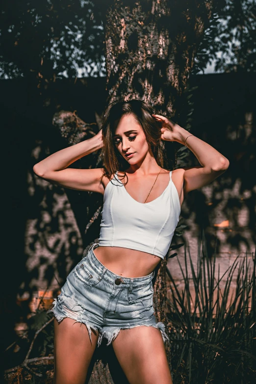 a beautiful young woman posing in front of a tree, by Robbie Trevino, trending on pexels, croptop and shorts, outfit : jeans and white vest, wearing : tanktop, best color graded