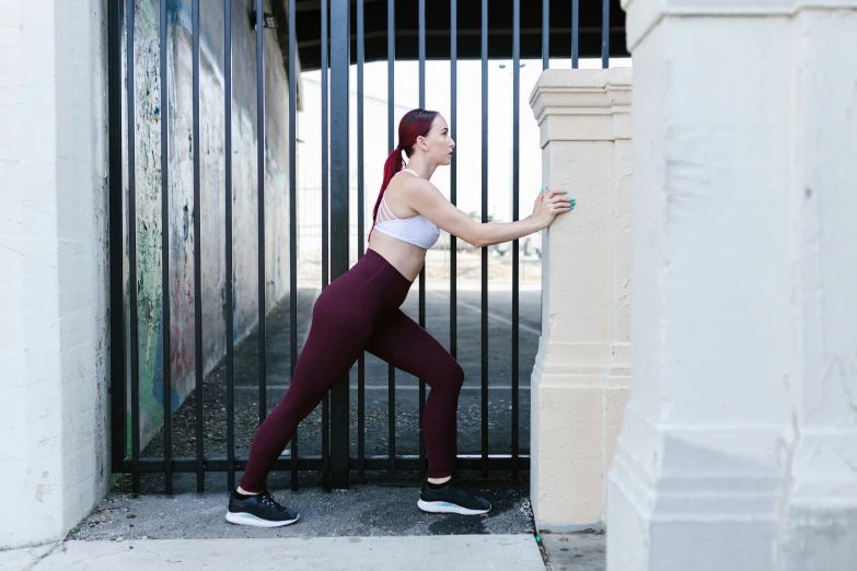 a woman in a sports bra top leaning against a gate, a photo, by Jacqui Morgan, pexels contest winner, happening, tights; on the street, maroon, waist reaching ponytail, pixeled stretching