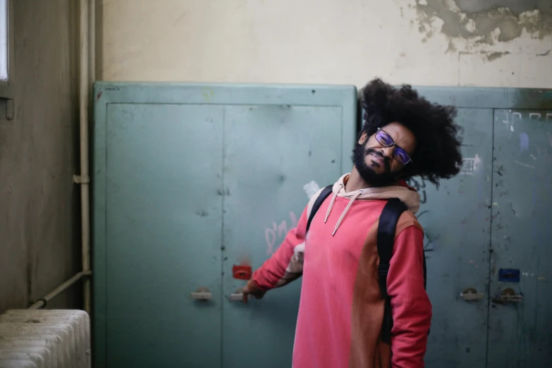 a man with a backpack standing in front of a door, an album cover, pexels, afro, bearded, pink, in a warehouse