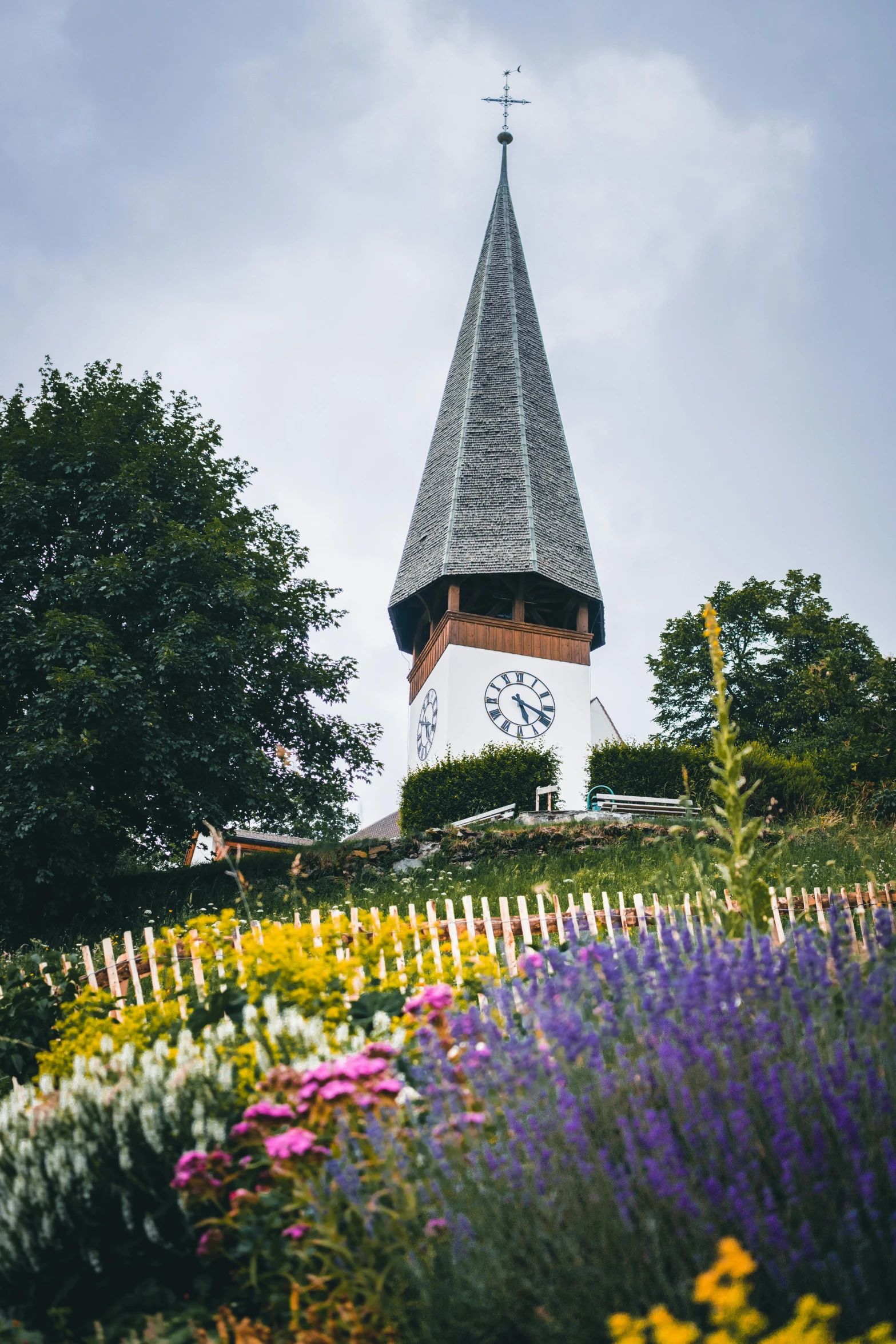 a clock tower sitting on top of a lush green hillside, inspired by Karl Stauffer-Bern, flower garden, lead - covered spire, nordic summer, cottagecore flower garden