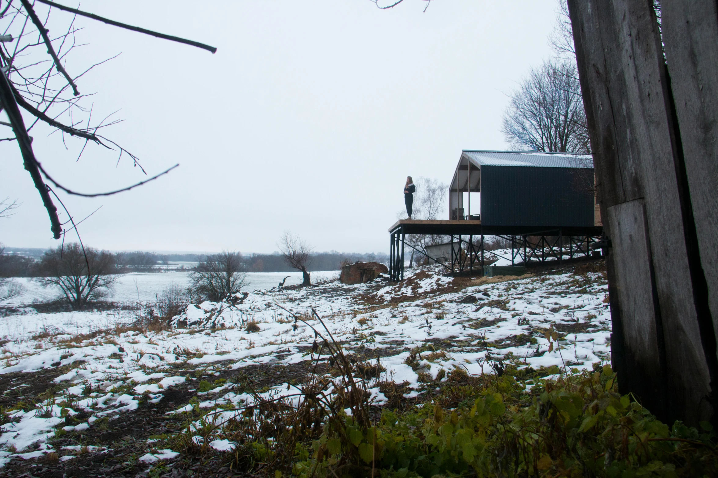 a couple of people standing on top of a snow covered field, by Jessie Algie, temporary art, building along a river, in a cabin, black steel buildings, on stilts