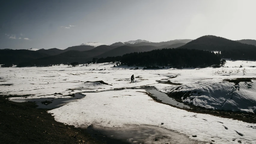 a person walking across a snow covered field, inspired by Kōno Michisei, unsplash contest winner, rocky mountains and a river, spring winter nature melted snow, dark and white, fishing